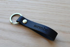 A black leather keychain strap with the name 'serenbe' engraved on it, attached to a gold-tone split ring and rivet, resting on a light wood surface.