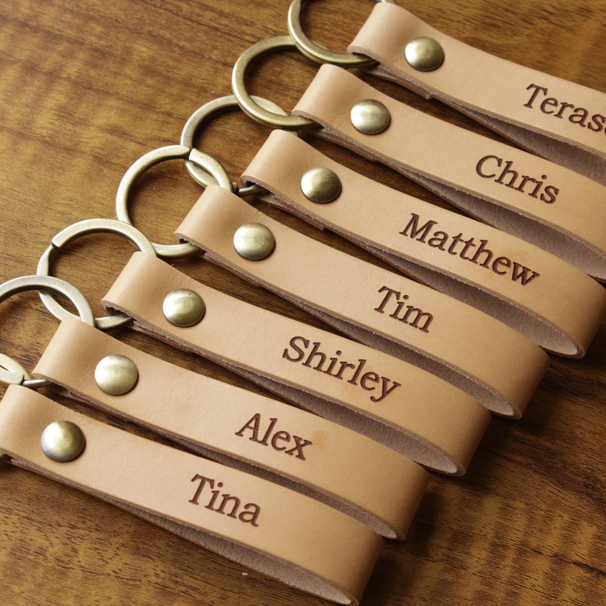Multiple leather keychains with names printed on them, laid out on a wooden surface.