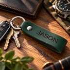 SANTJAG personalized Forest Green leather keychain laser engraved with the name JASON, displayed with metal keys, a wristwatch and a leather wallet on a wooden desk.