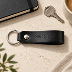 Black leather keychain engraved with John 10:10 and a cross, with silver hardware beside a metal key and black notebook.