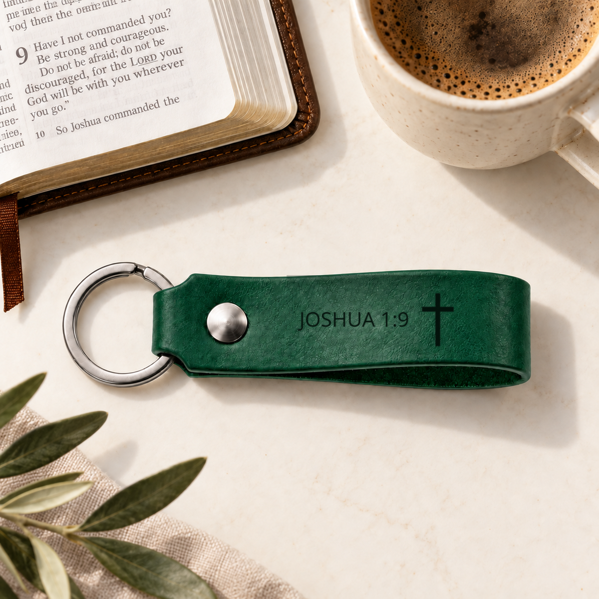 Green leather keychain engraved with Joshua 1:9 and a cross, with silver hardware beside an open Bible.