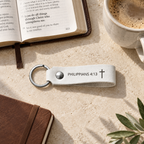 White leather keychain engraved with Philippians 4:13 and a cross, displayed beside an open Bible, coffee and a brown notebook.