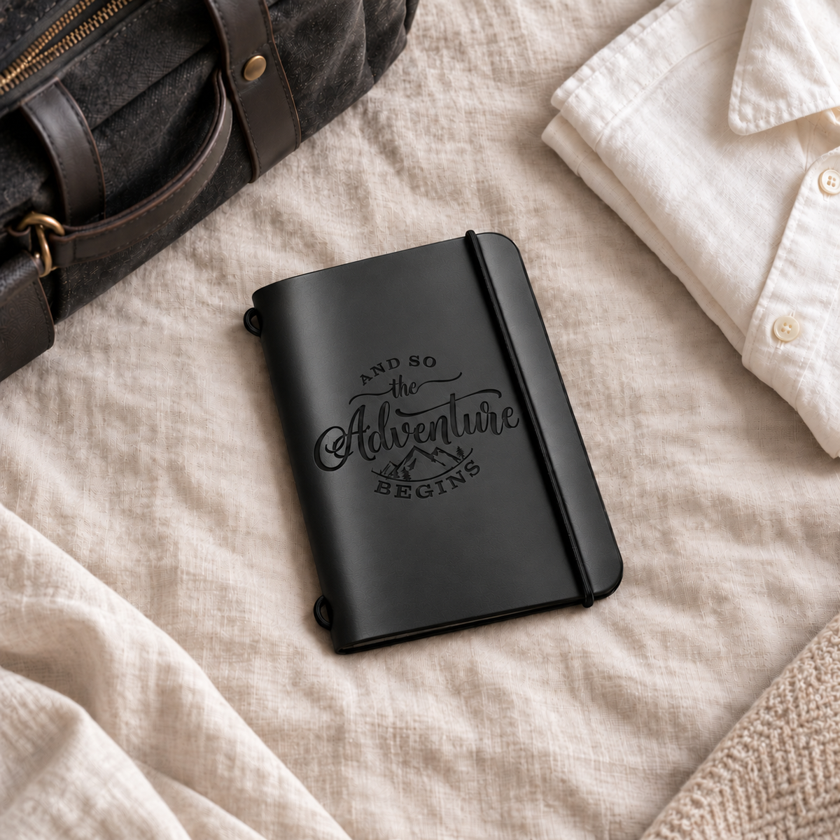 SANTJAG smooth black leather passport holder engraved with “And So the Adventure Begins” and mountains, resting on beige linen beside a travel bag and folded white shirt.