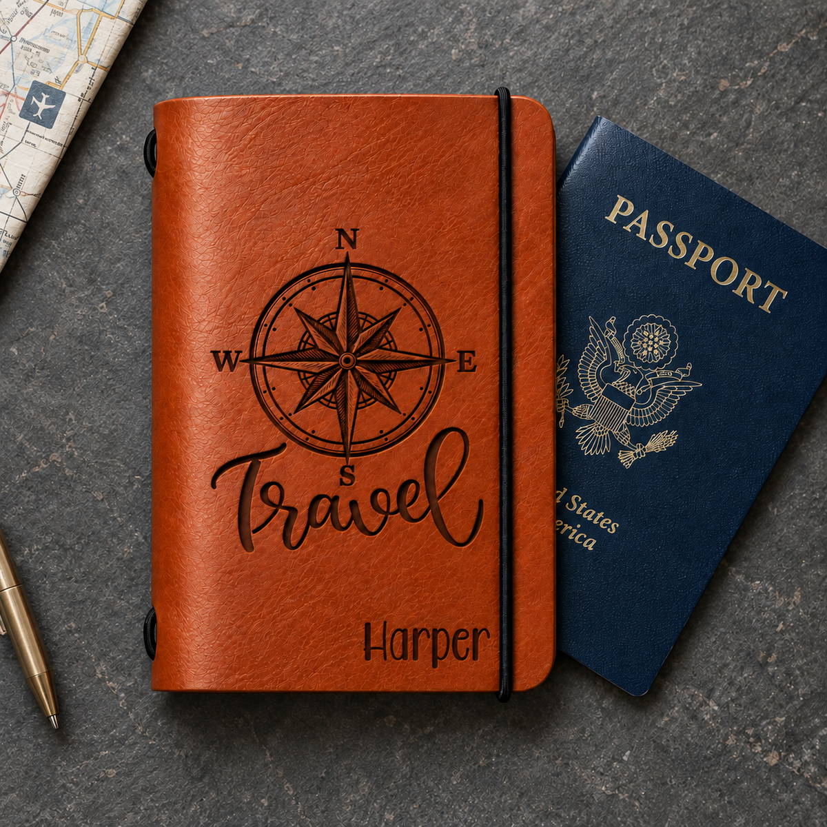 SANTJAG brown leather passport holder engraved with a compass rose, “Travel,” and the name Harper, beside a blue passport on a dark stone surface.