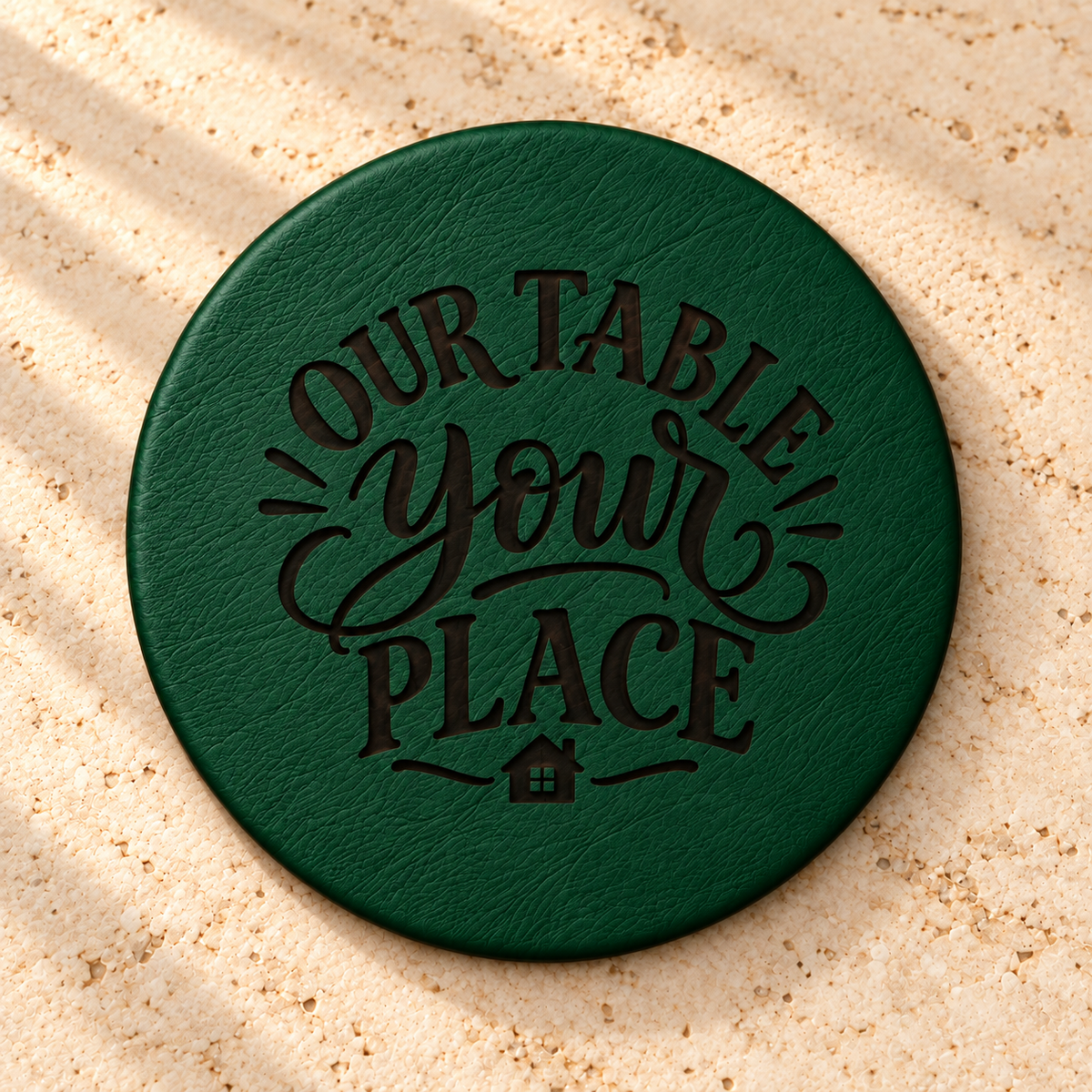 Green round leather coaster engraved with “Our Table Your Place” and a small house design on a light stone surface.