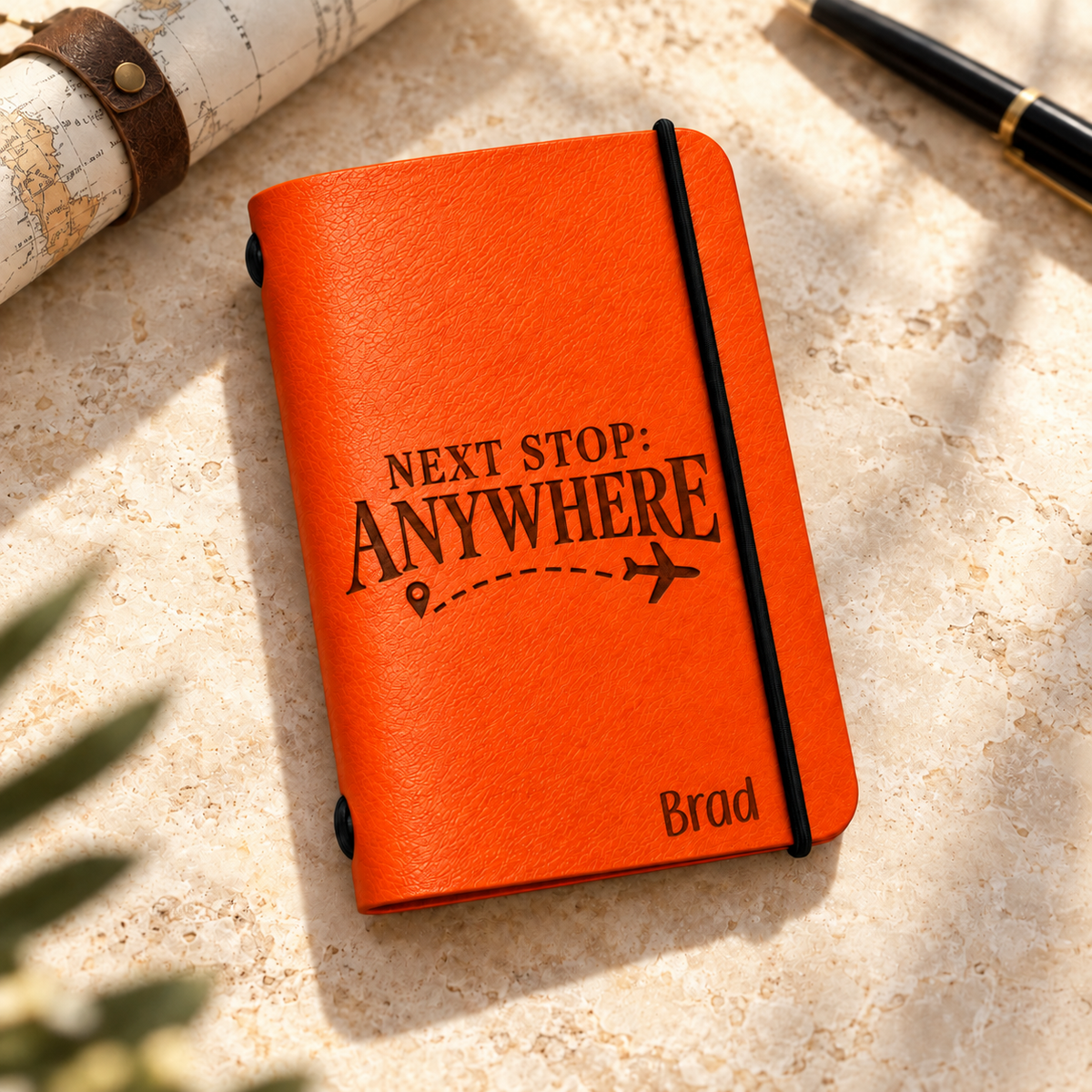 SANTJAG orange leather passport holder engraved with “Next Stop: Anywhere,” an airplane, and the name Brad, on light stone beside a rolled map and pen.