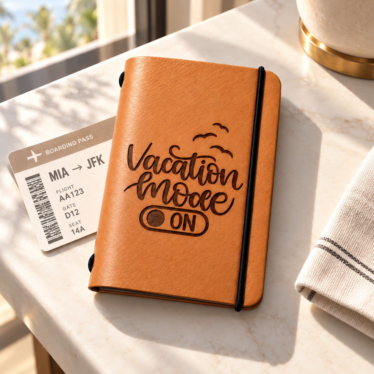 SANTJAG tan leather passport holder engraved with “Vacation Mode On” and flying birds, featuring a black elastic closure, beside a boarding pass on a sunlit marble table.