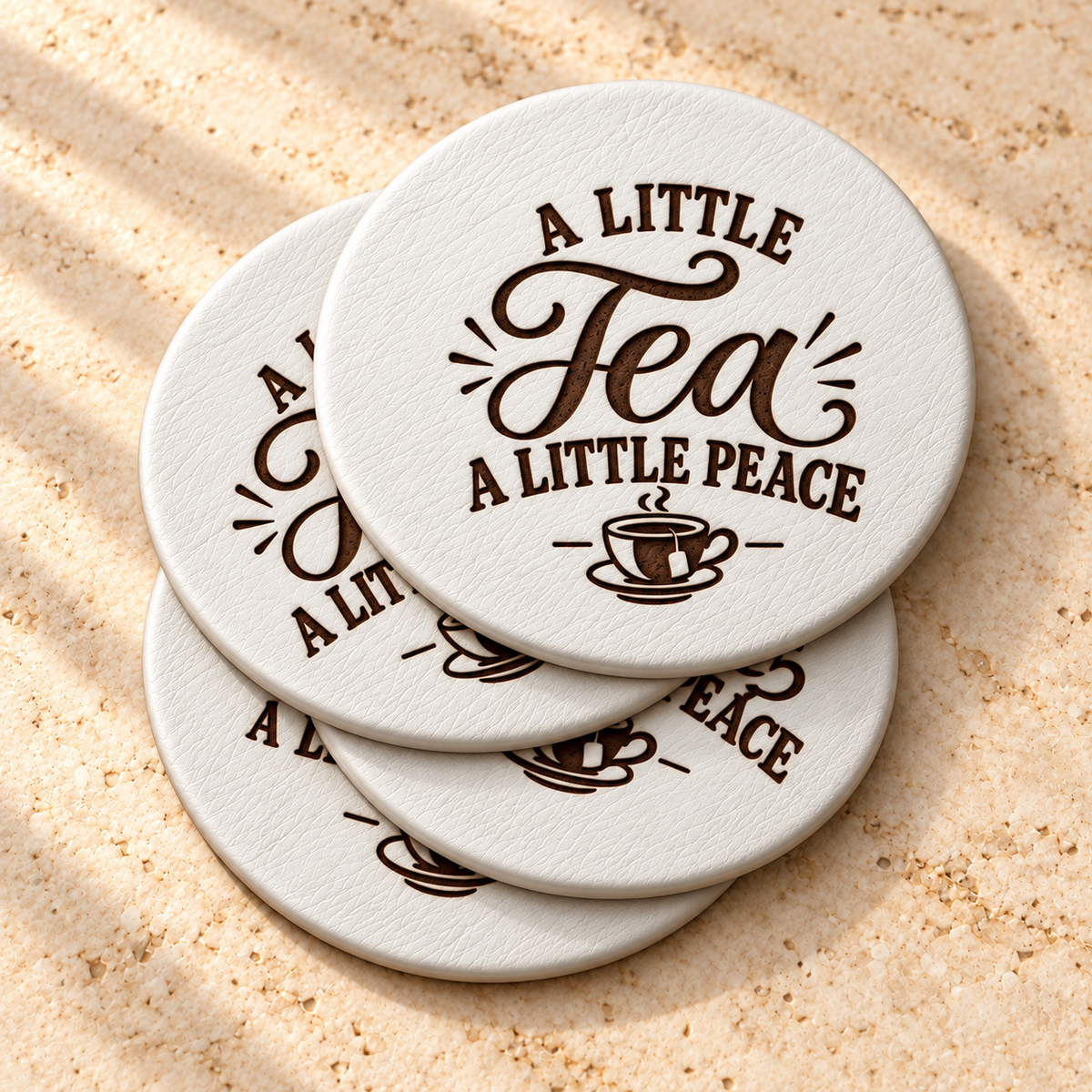 Stacked white round leather coasters engraved with “A Little Tea A Little Peace” and a teacup design on a light stone surface.