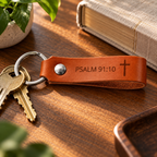Close-up of a cognac leather keychain engraved with Psalm 91:10 and a cross, attached to keys on a wooden table.