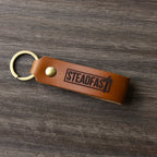 A brown leather keychain with the word 'Steadfast' printed on it, placed on a wooden surface.