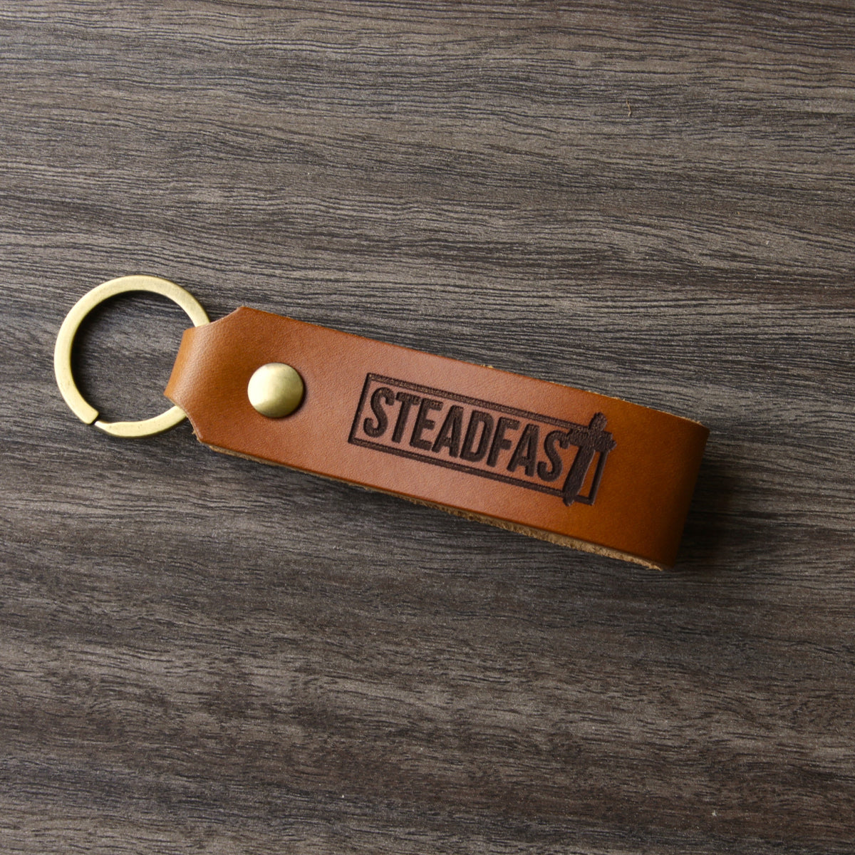 A brown leather keychain with the word 'Steadfast' printed on it, placed on a wooden surface.